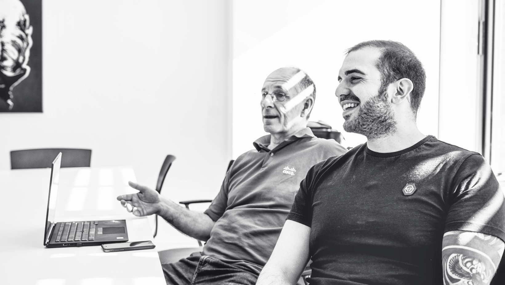 Zwei Männer sitzen an einem Tisch in einem hellen Raum, lächeln und unterhalten sich. Ein Mann gestikuliert mit der Hand, während der andere lacht. Auf dem Tisch stehen ein Laptop und ein Telefon. Die Atmosphäre wirkt entspannt und freundlich.