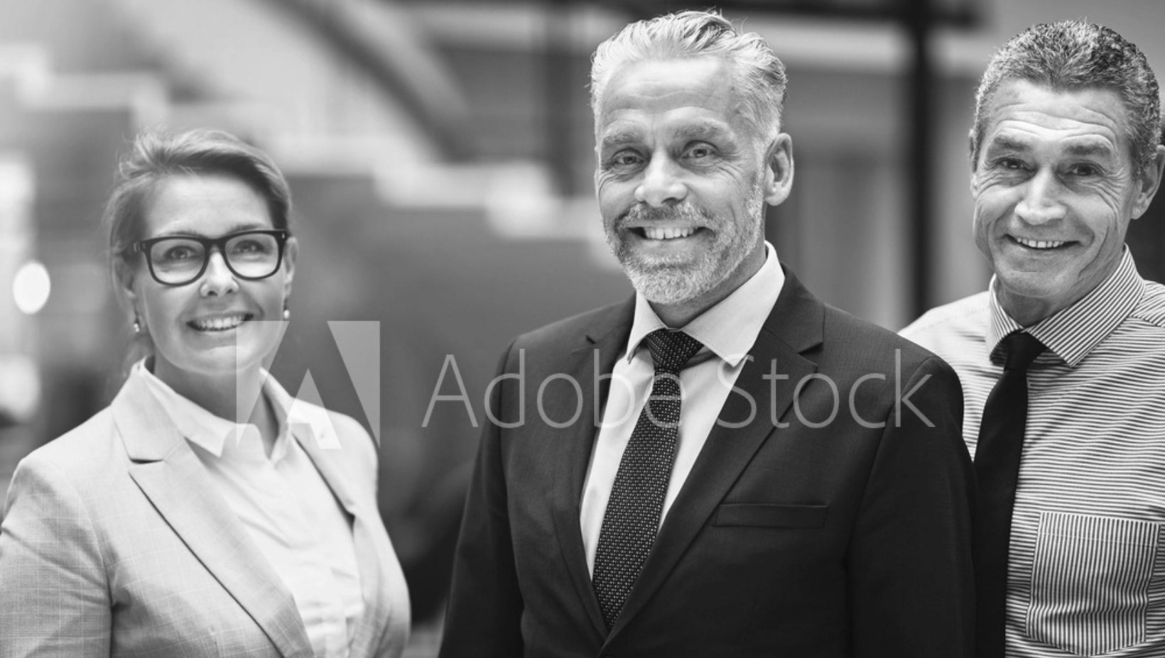 Drei professionell gekleidete Erwachsene, zwei Männer und eine Frau, stehen lächelnd in einer modernen Büroumgebung zusammen. Das Bild ist in Schwarz-Weiß und mit einem Adobe Stock-Wasserzeichen versehen.