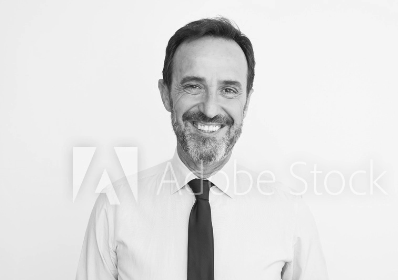 Schwarz-Weiß-Foto eines lächelnden Mannes mittleren Alters mit Bart, der ein helles Hemd und eine dunkle Krawatte trägt und vor einem schlichten weißen Hintergrund steht.