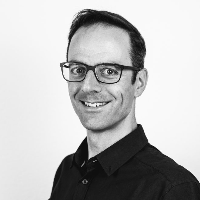 Ein Mann mit kurzen dunklen Haaren und Brille lächelt in die Kamera. Er trägt ein dunkles Button-up-Hemd und ist auf einem Schwarz-Weiß-Foto vor einem schlichten, hellen Hintergrund abgebildet.