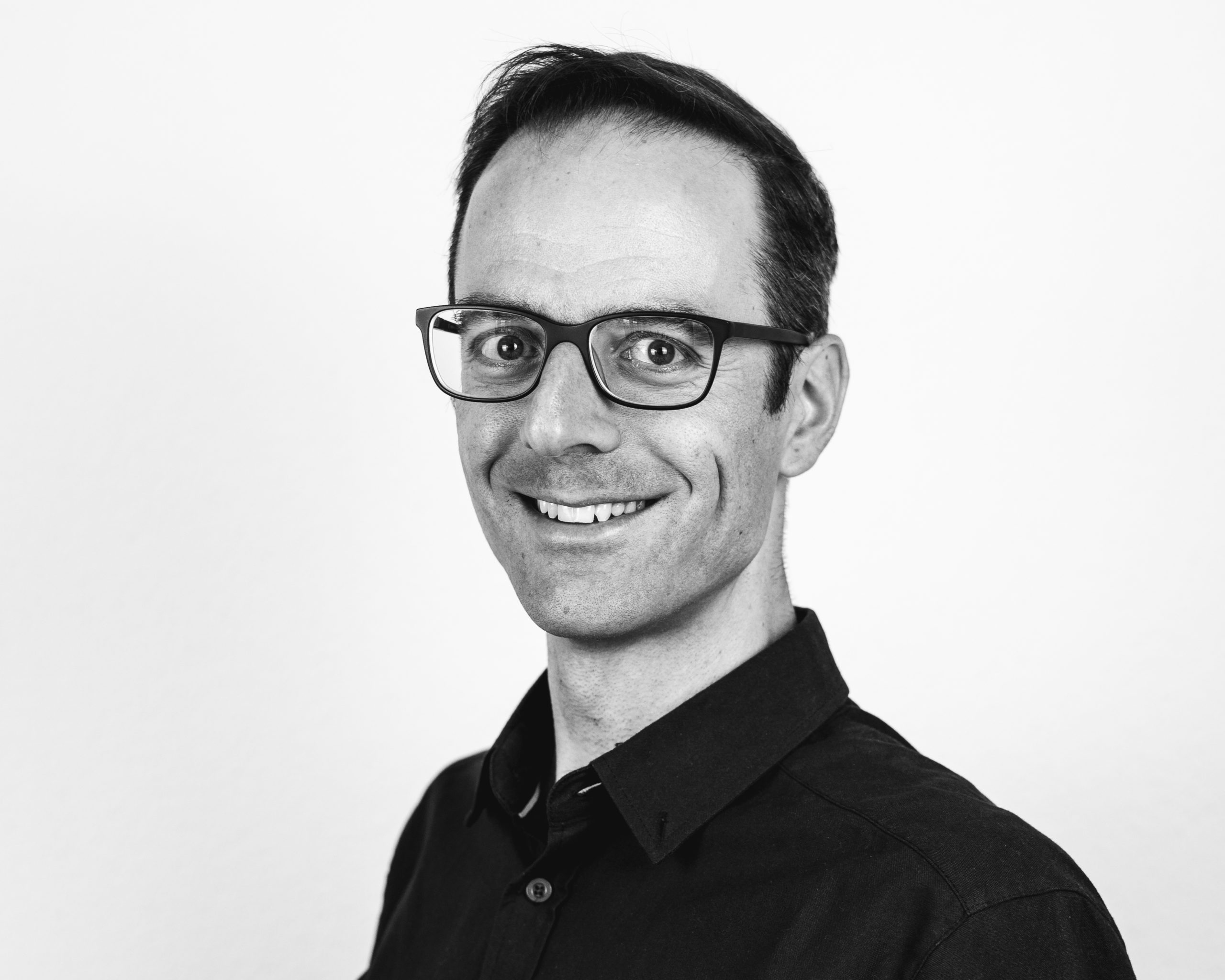 A man with short dark hair and glasses smiles at the camera. He is wearing a dark collared shirt. The image is black and white with a plain light background.