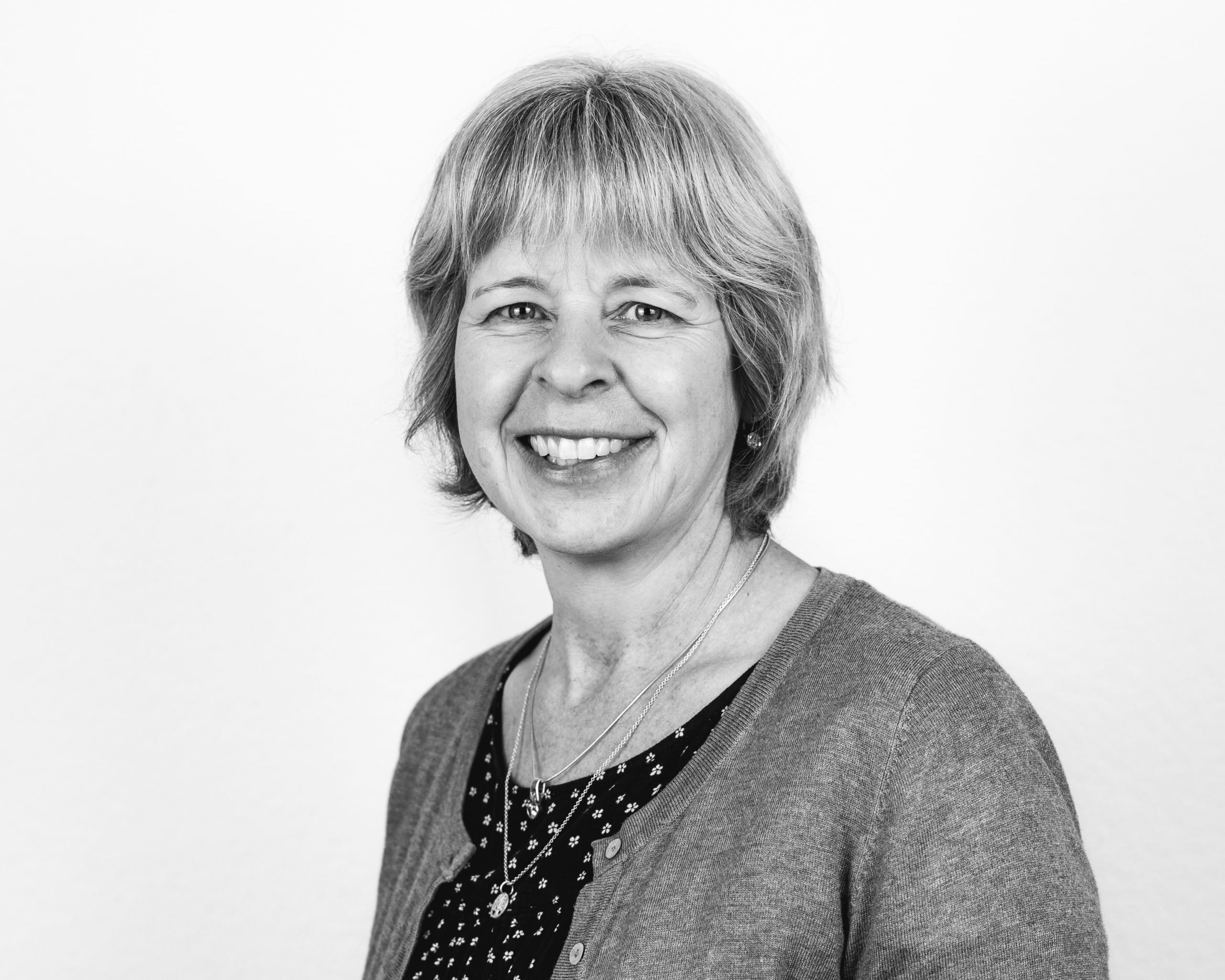 Black and white portrait of a middle-aged woman with short, light hair, smiling, wearing a patterned blouse, a cardigan, and a necklace, standing against a plain background.