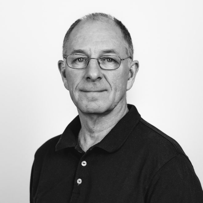 Ein Mann mittleren Alters mit kurzen Haaren und Brille, der ein dunkles Hemd mit Kragen trägt, posiert vor einem schlichten, hellen Hintergrund. Das Bild ist in schwarz-weiß gehalten.