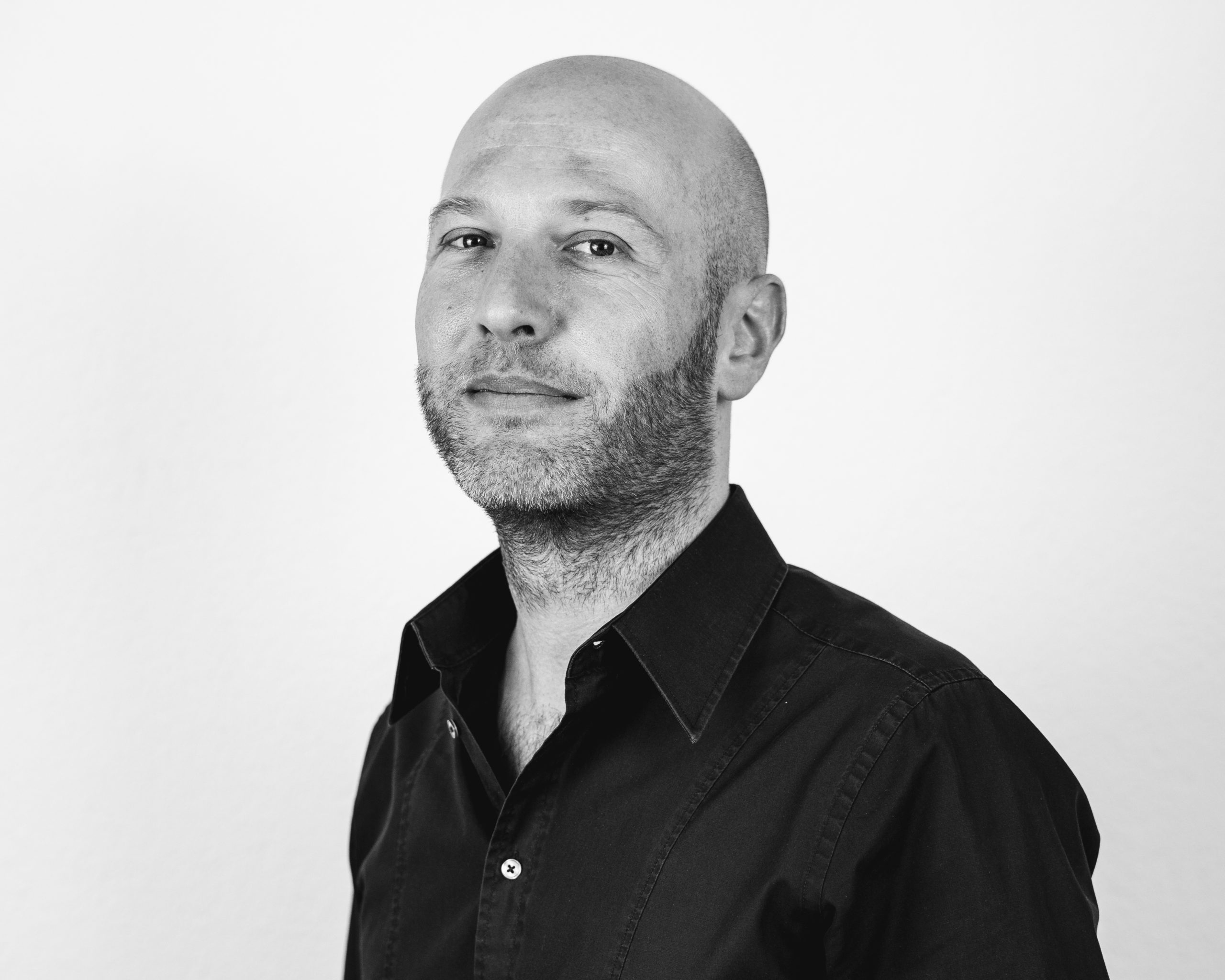 A bald man with stubble wearing a dark button-up shirt looks confidently at the camera against a plain, light background in a black-and-white portrait.