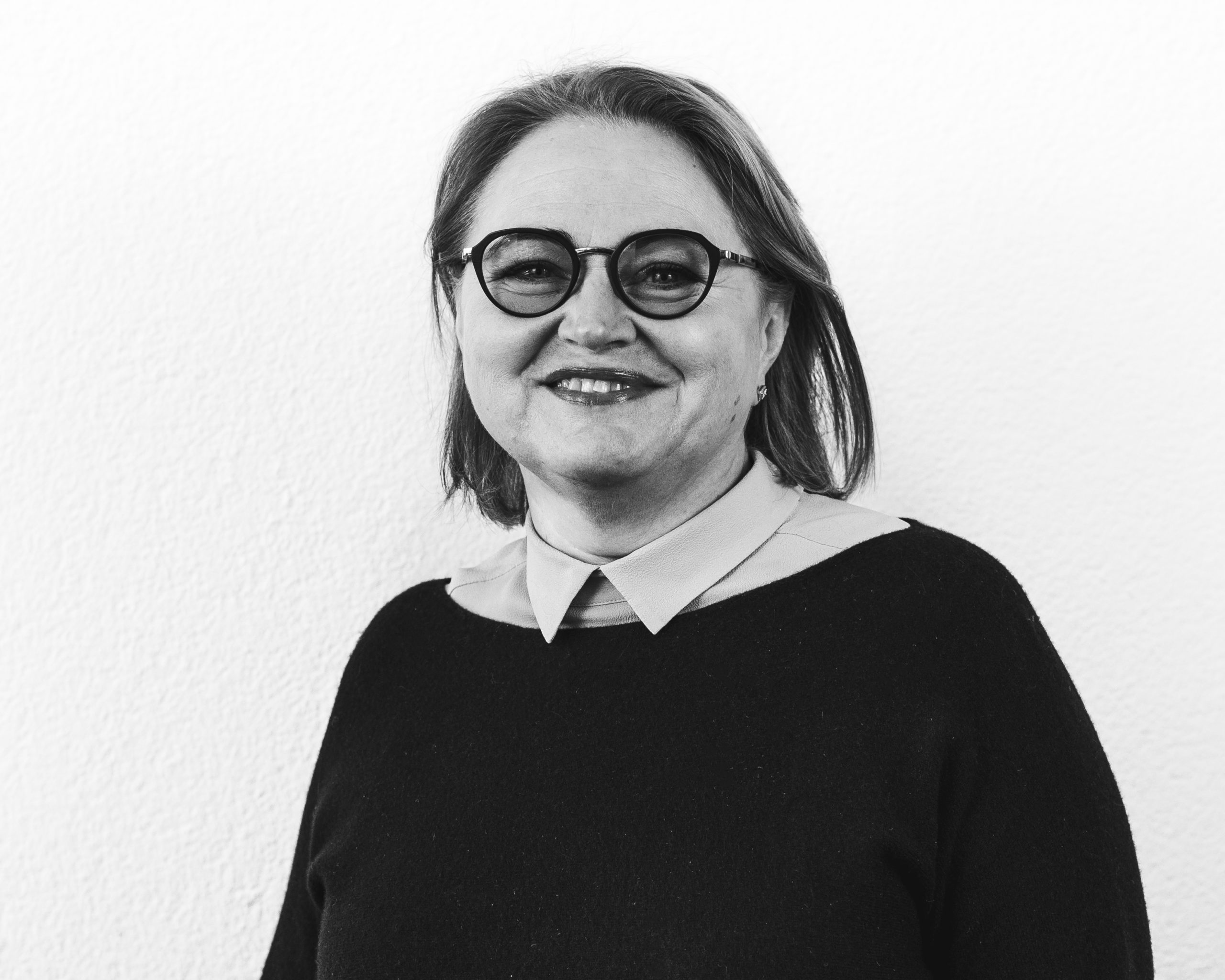 A woman with shoulder-length hair and glasses smiles at the camera. She is wearing a collared shirt under a dark sweater. The photo is in black and white with a plain background.