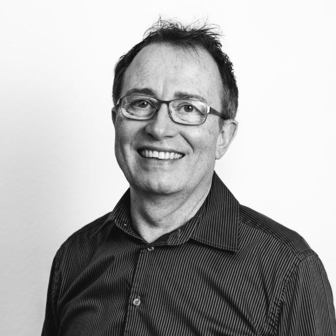 Ein Mann mit Brille und gestreiftem, dunklem Button-up-Hemd lächelt in die Kamera. Das Foto ist schwarz-weiß mit einem schlichten hellen Hintergrund.