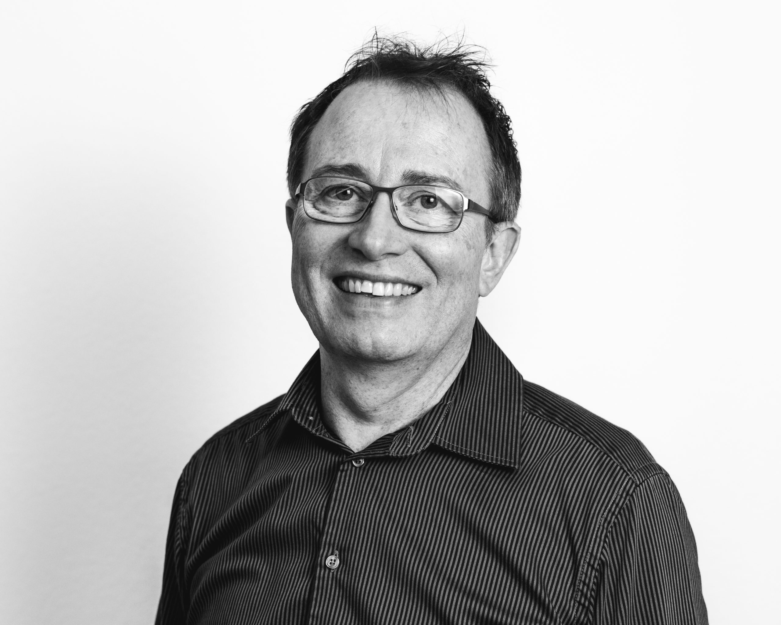 A man with glasses and short hair smiles at the camera. He is wearing a striped, button-up shirt, and the photo is in black and white with a plain background.