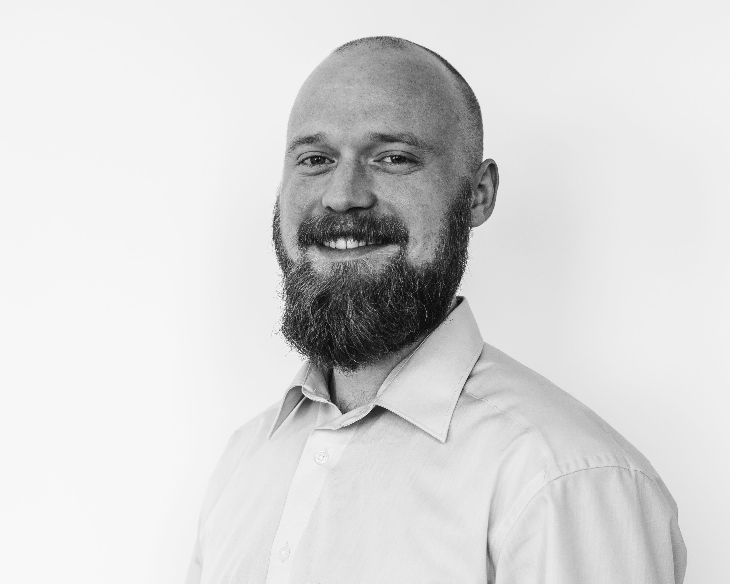 A bearded man with a shaved head, wearing a light-colored button-up shirt, smiles at the camera against a plain white background. The photo is in black and white.