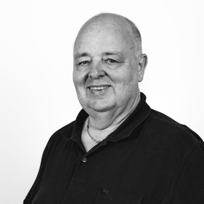 Ein lächelnder Mann mittleren Alters mit Glatze und dunklem Poloshirt steht vor einem schlichten weißen Hintergrund. Das Foto ist in schwarz-weiß gehalten.