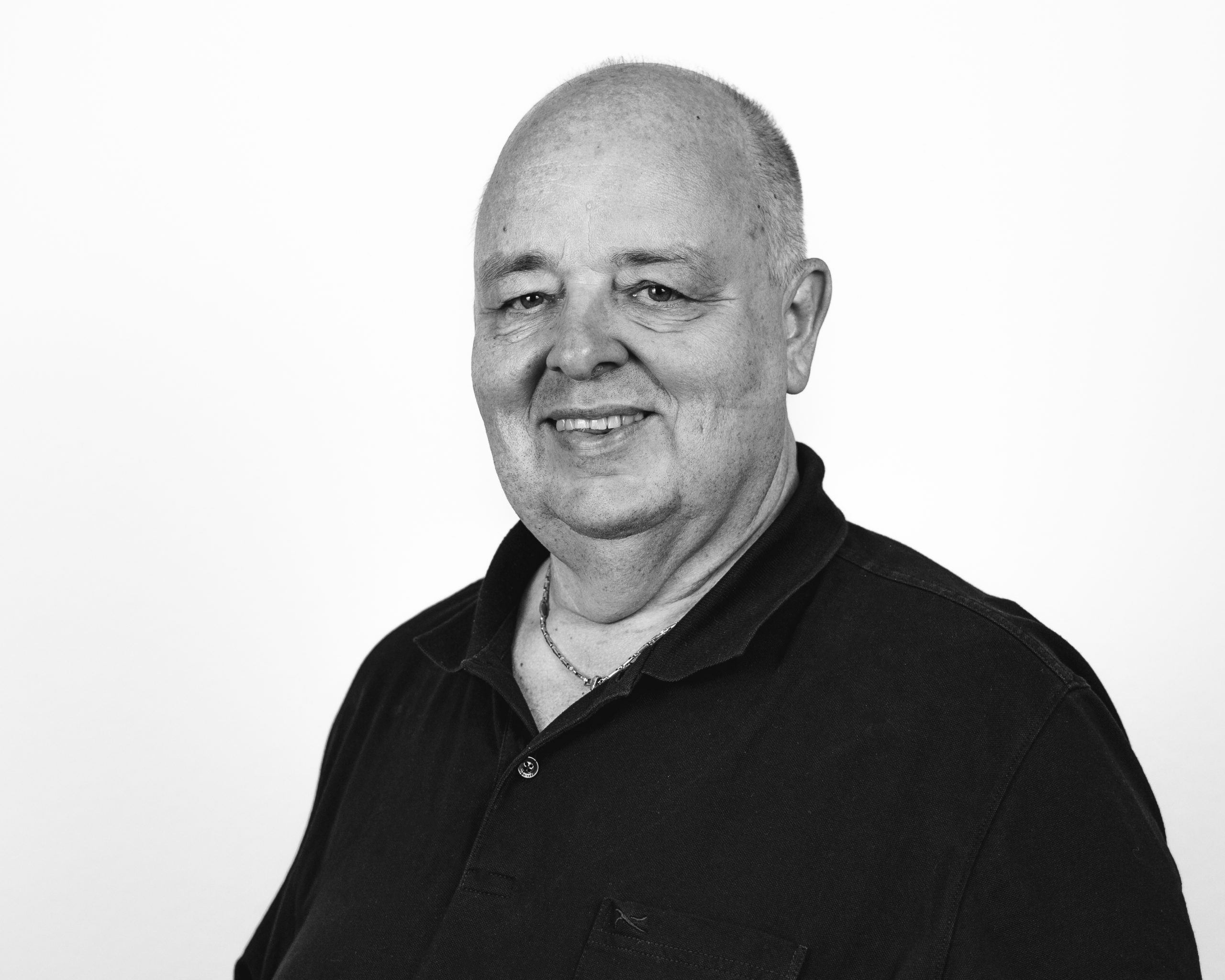 A smiling older man with a bald head wearing a dark collared shirt, photographed against a plain white background. The image is in black and white.