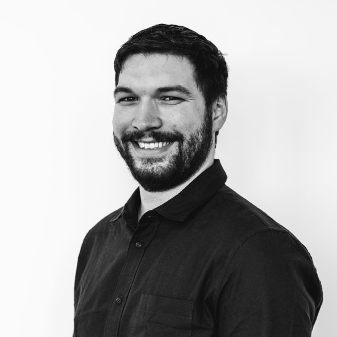 Ein Mann mit kurzen dunklen Haaren und einem Bart lächelt in die Kamera. Er trägt ein dunkles Button-up-Hemd und steht vor einem schlichten weißen Hintergrund. Das Foto ist in schwarz-weiß gehalten.