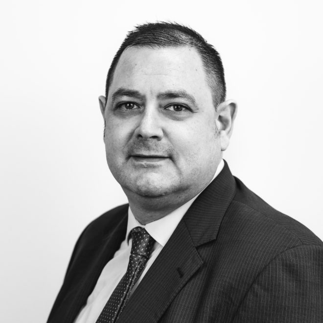 Schwarz-Weiß-Porträt eines Mannes in Anzug und Krawatte, der mit neutralem Blick in die Kamera schaut, vor einem schlichten hellen Hintergrund.