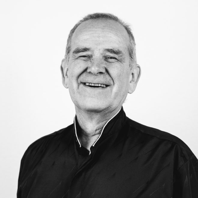 Schwarz-Weiß-Foto eines älteren Mannes, der freundlich in die Kamera lächelt. Er hat kurze Haare und trägt ein dunkles, geknöpftes Hemd. Der Hintergrund ist schlicht und hell.