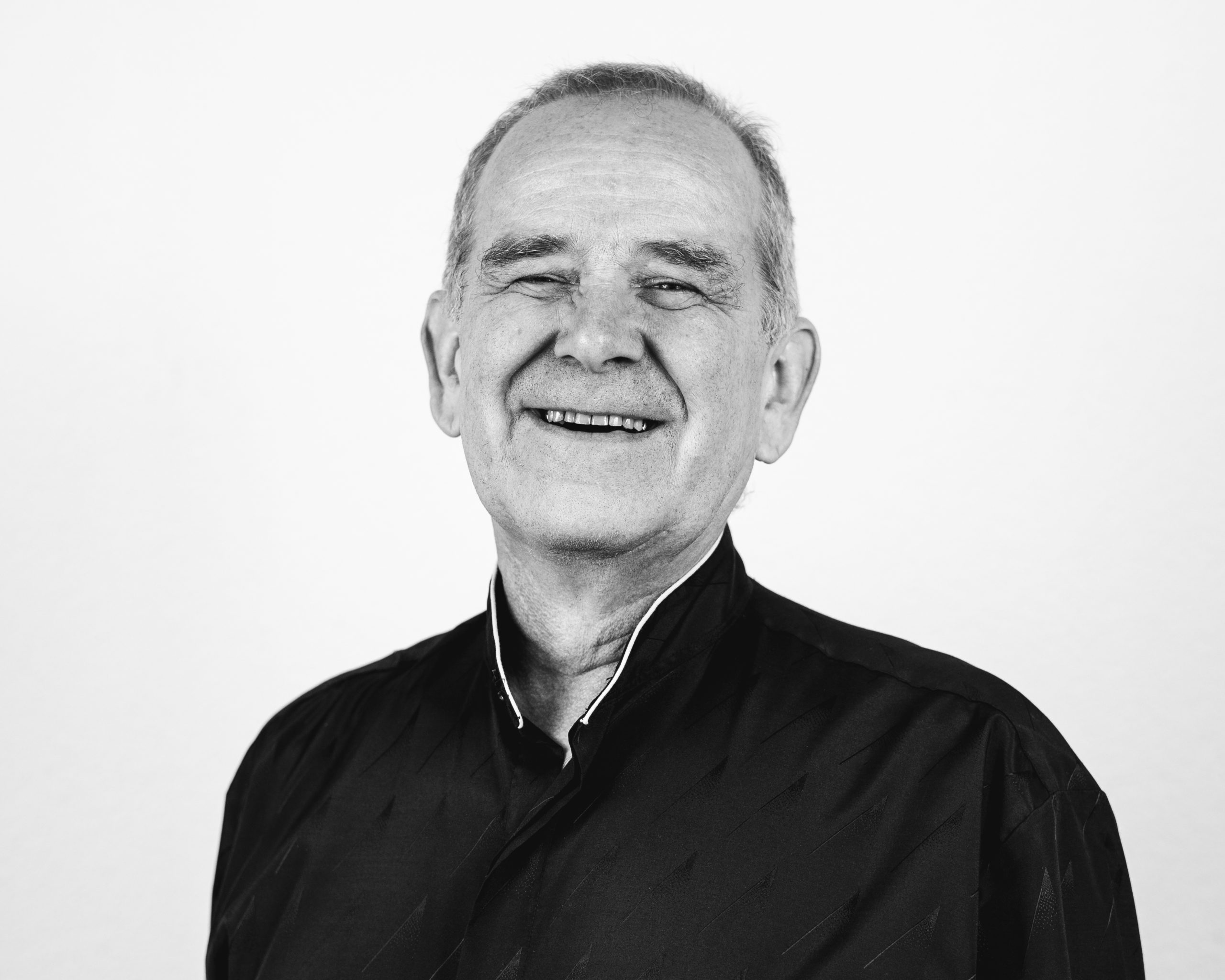 An older man with short gray hair smiles warmly at the camera. He is wearing a dark, collared shirt and is posed against a plain, light-colored background. The photo is in black and white.