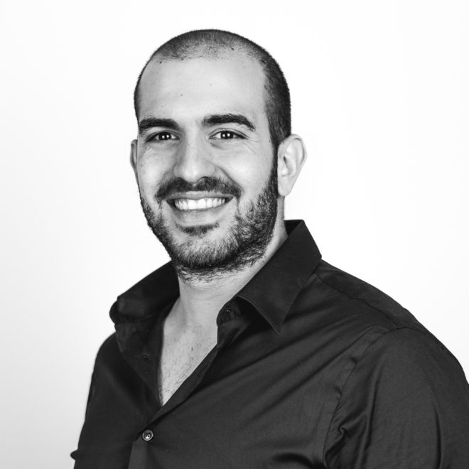 Ein Mann mit kurzen Haaren und Bart, der ein dunkles Button-up-Hemd trägt, lächelt in die Kamera vor einem schlichten weißen Hintergrund. Das Foto ist schwarz-weiß.