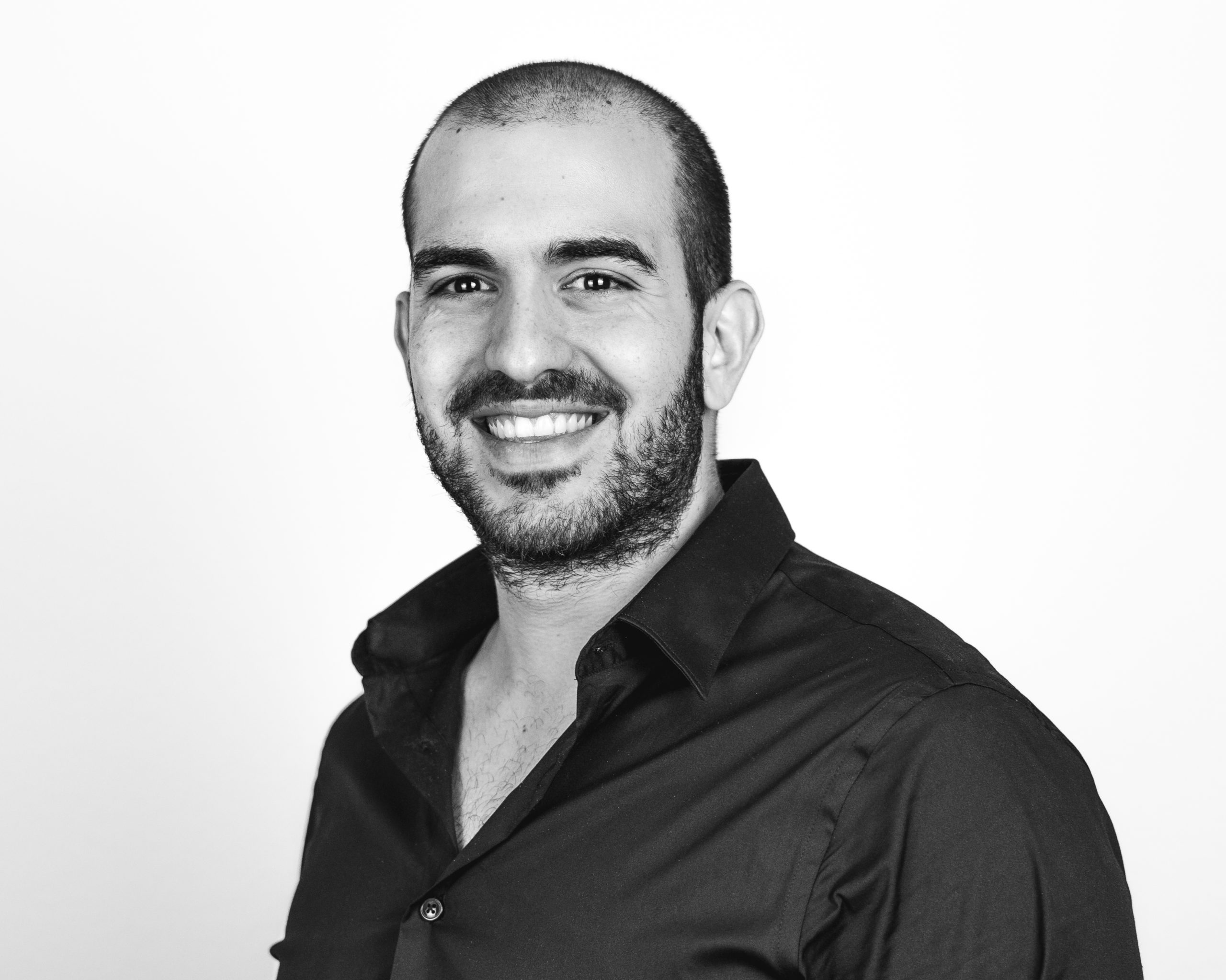 A man with a shaved head and beard, wearing a dark button-up shirt, smiles at the camera against a plain white background.