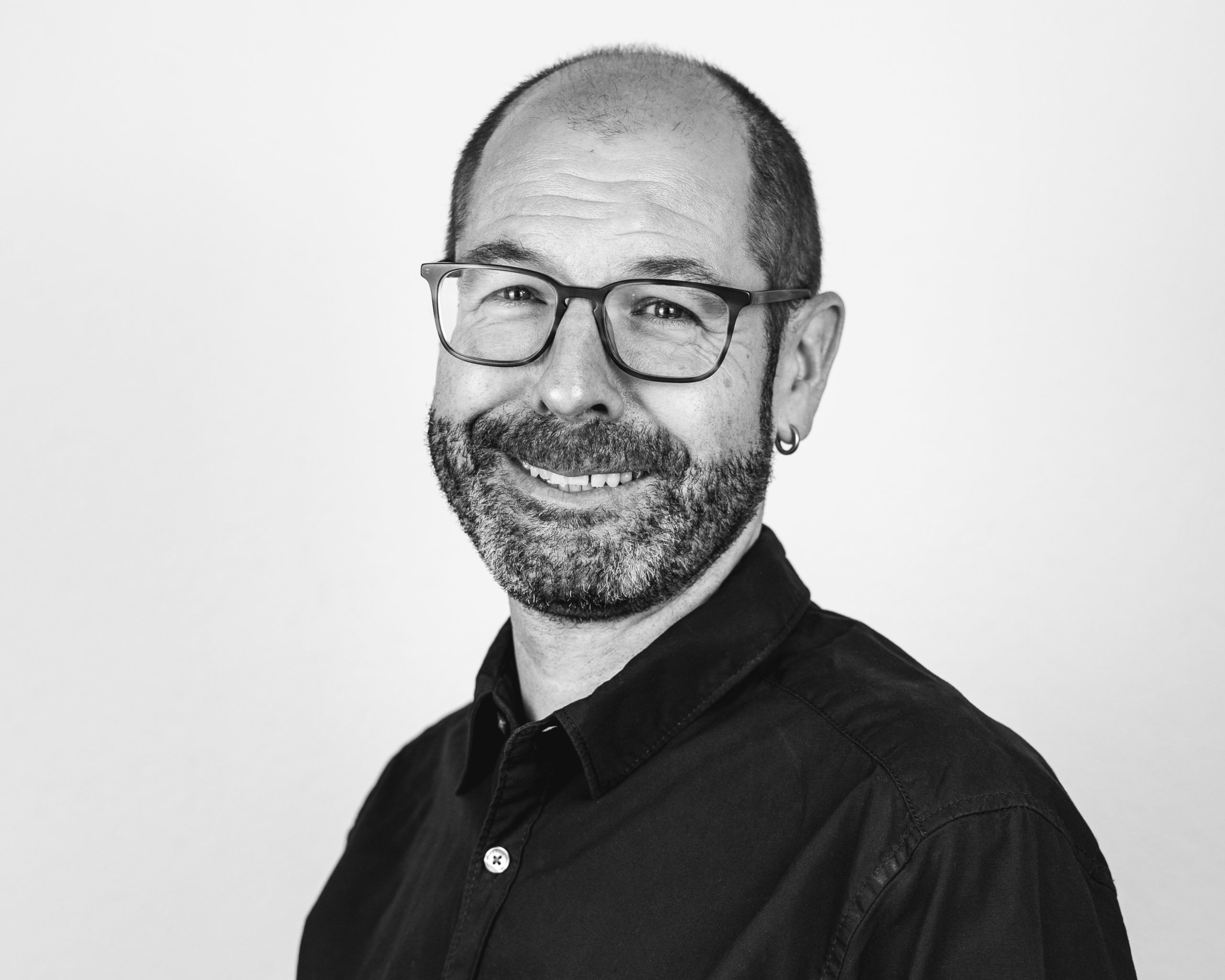 A smiling middle-aged man with glasses, a beard, and an earring, wearing a dark button-up shirt, poses against a plain light background in a black-and-white photo.
