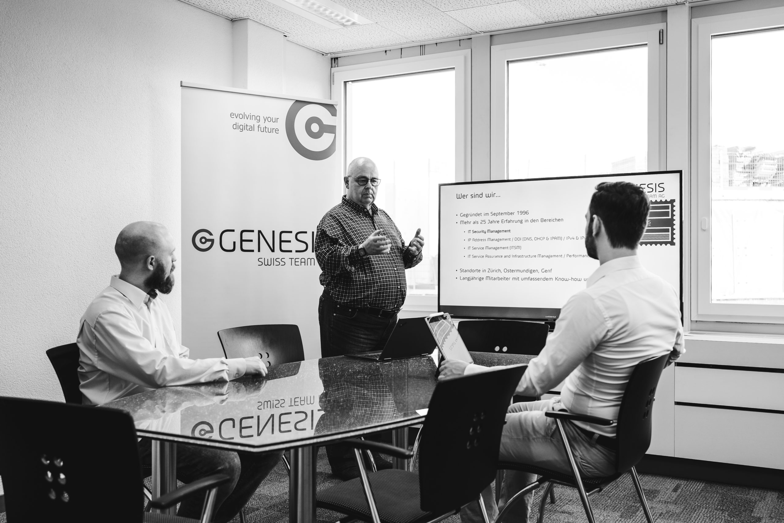 Drei Männer in Geschäftskleidung befinden sich in einem modernen Besprechungsraum. Einer steht und präsentiert Informationen auf einem Bildschirm, während die beiden anderen an einem Glastisch sitzen und aufmerksam zuhören. Ein GENESIS-Banner ist zu sehen.