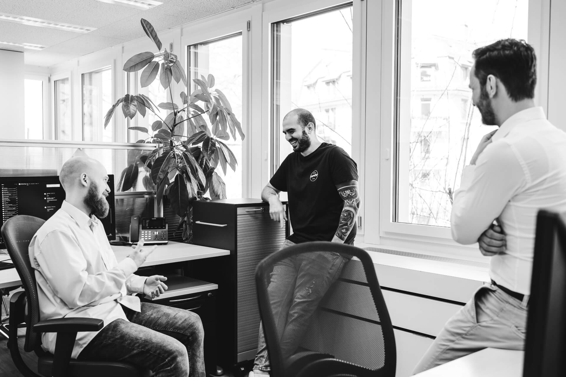 Drei Männer führen ein lockeres Gespräch in einem modernen Büro. Ein Mann sitzt an einem Schreibtisch, ein anderer lehnt an einem Schrank, und der dritte steht in der Nähe. Im Hintergrund sind große Fenster und Zimmerpflanzen zu sehen.